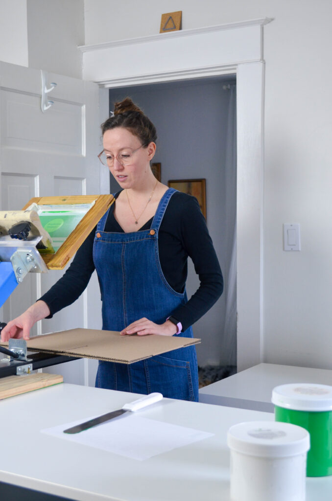 A woman wearing a denim jumper screen prints a logo on a box.