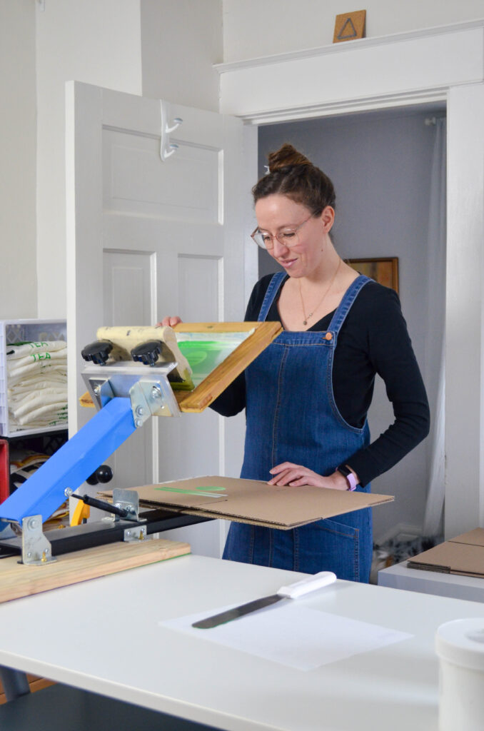 A woman wearing a denim jumper screen prints a logo on a box.