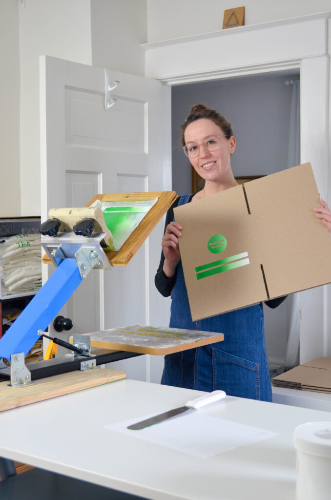 A woman wearing a denim jumper screen prints a logo on a box.