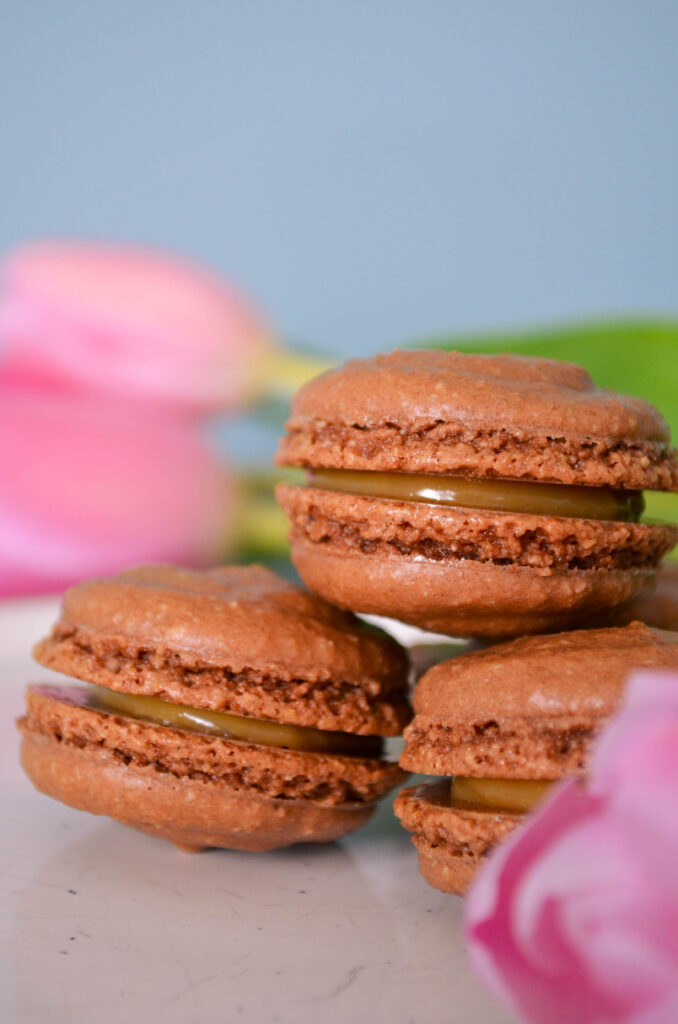 Chocolate macarons filled with dulce de leche, surrounded by pink tulips.
