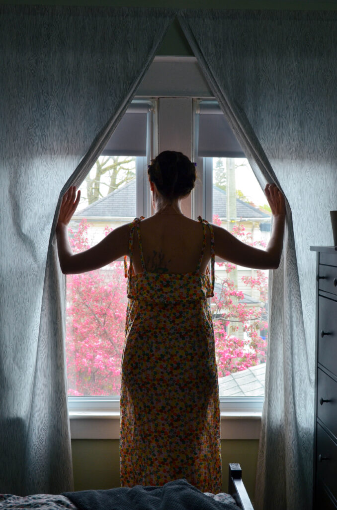 A woman in a nightgown, standing in front of a window, opening the curtains.